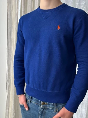 Clean blå Ralph Lauren sweatshirt, storlek S - Clean Blå Ralph Lauren sweatshirt med orange logga i 84% bomull och 16% polyester  Mycket bra skick, sömmen har släppt lite i nacken men absolut inget som påverkar användningen   Storlek S