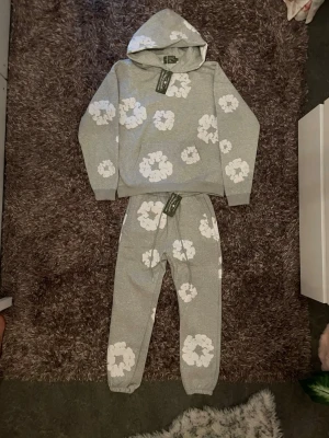 Denim tears  - Mysigt grått loungewear-set med hoodie och byxor, dekorerat med stora vita blommor över hela plagget. Hoodien har luva och ribbade muddar, byxorna har resår i midjan och benslut. Perfekt för chill dagar hemma. Förpackning finns med!