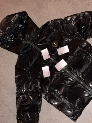 Svart pufferjacka från Trapstar Irongate  - Snygg svart pufferjacka från Trapstar med glansig finish och stor huva. Jackan har dragkedja framtill och Trapstar-logga broderad på ryggen. Perfekt för kalla dagar och riktigt street-inspirerad stil.