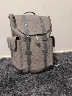 Grå Louis Vuitton ryggsäck i läder - Säljer en grå ryggsäck från Louis Vuitton i präglat läder med klassiskt monogrammönster. Ryggsäcken har två sidofickor med lock, justerbara axelband och snygga detaljer i mörkgrått. Perfekt för dig som vill ha en exklusiv och stilren väska.
