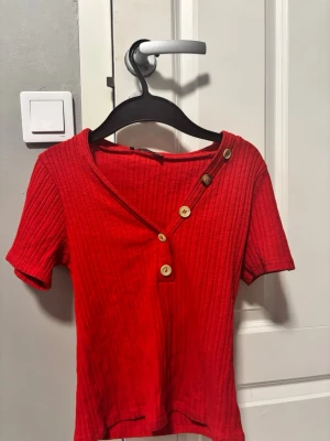 Röd ribbad topp med knappar köpt utomlands  - Snygg röd ribbad topp med korta ärmar och unik v-ringning dekorerad med fyra träknappar. Toppen har en croppad passform. Perfekt för dig som vill sticka ut med en cool detalj.
