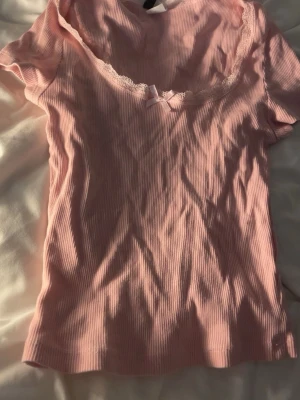 Rosa utringad topp med spetskant - Supersöt ljusrosa utringad topp med korta ärmar. Urringningen är dekorerad med en liten rosett och spetskant som ger en gullig vibe. Perfekt att styla med jeans eller kjol för en trendig look. Materialet känns mjukt och stretchigt.