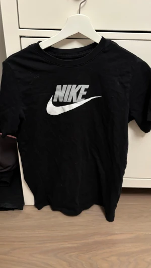 Svart Nike t-shirt med logga - Klassisk svart t-shirt från Nike med stor vit logga på bröstet. T-shirten har rund hals och korta ärmar, perfekt för en sportig och avslappnad stil. Tillverkad i mjuk bomull för skön komfort.