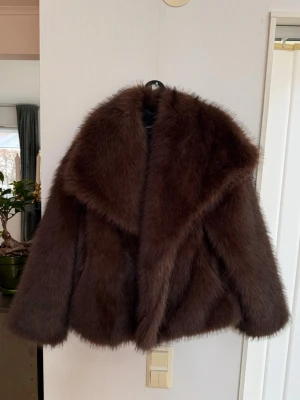 Mörkbrun fluffig pälsjacka - Säljer en mörkbrun pälsjacka med oversized krage och breda ärmar. Jackan har en lyxig, fluffig känsla och ger en riktigt cool vintage-vibe. Perfekt för dig som vill sticka ut och hålla dig varm under kalla dagar. Materialet är fuskpäls.