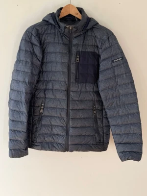 Mörkblå dunjacka från Calvin Klein - Snygg mörkblå dunjacka från Calvin Klein med huva och quiltad design. Jackan har en svart bröstficka med dragkedja, två sidofickor med dragkedja och diskret logga på ärmen. Perfekt för kalla dagar och riktigt stilren look.