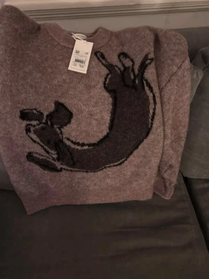 Brun stickad tröja med hundmotiv - Stickad tröja från Carin Wester  i brun  med svart hundmotiv på framsidan. Tillverkad i en mjuk mix av polyamid, ull, alpacka och elastan. Rund halsringning och avslappnad passform, perfekt för dig som gillar unika detaljer.