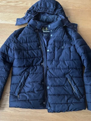 Svart pufferjacka från Barbour - Snygg svart pufferjacka från Barbour med huva och dragkedja. Jackan har två snedställda fickor med dragkedja, bröstficka med knapp och quiltad design. Perfekt för kalla vinterdagar och har en klassisk look med diskreta detaljer.