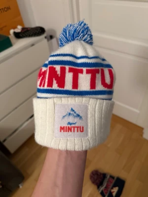 Vit och blå Minttu mössa med tofs - Säljer en vit ribbstickad Minttu mössa med blå ränder och röd text. Mössan har en blåvit tofs på toppen och en uppvikt kant med Minttu-logga framtill. Perfekt för kalla dagar 