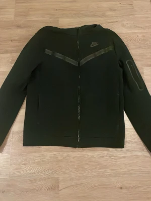 Svart Nike hoodie med dragkedja - Svart hoodie från Nike med hel dragkedja framtill och diskreta svarta detaljer över bröstet. Tröjan har en huva och lång ärm, samt en liten ficka på ena ärmen. Materialet känns mjukt och sportigt, perfekt för en avslappnad stil.köpt för 1200 kr. I storleken passar den personer som har S. Bara använd ett par gånger