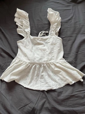 Vit volangblus med broderi och knyt - Supersöt vit blus med volangärmar och broderade detaljer. Toppen har knyt i ryggen och en lätt utsvängd nederdel. Perfekt för dig som gillar romantisk och feminin stil. Materialet känns som bomull och toppen är ärmlös med breda axelband.