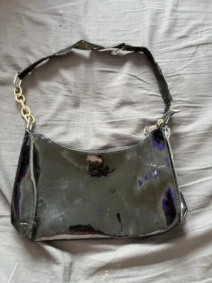 Svart lack axelväska med kedjedetalj - Snygg svart axelväska i glansigt lackmaterial med silvriga kedjedetaljer på axelremmen. Väskan har en enkel, avlång form och stängs med dragkedja upptill. Perfekt för dig som gillar en trendig och edgy look. 