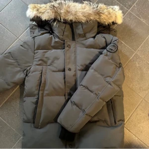 Canada goose  - Snygg svart dunjacka från Canada Goose med stor pälskantad huva och patch på ärmen. Jackan har dragkedja och knappar framtill, flera fickor och quiltad design. Perfekt för kalla vinterdagar och riktigt varm.