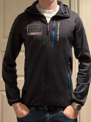 Svart vindjacka från Nautic XPRNC - Svart vindjacka med huva från Nautic XPRNC. Jackan har blå dragkedjor, tryck på bröstet och ärmen samt texten 'OFFSHORE RACING RS6G' och 'NAUTIC XPRNC RS6G'. Praktiska fickor och sportig design i ett slitstarkt material.