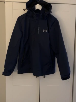 Blå vindjacka från Under Armour - Snygg blå vindjacka från Under Armour med huva och dragkedja framtill. Jackan har två dragkedjeförsedda fickor på sidorna och en diskret logga på bröstet. Perfekt för blåsiga dagar och sportiga outfits. Den är Vattentätt med goretex