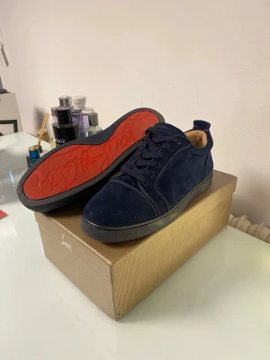 Christian Louboutin - Snygga blåa sneakers från Christian Louboutin med klassisk röd sula och signatur på sidan. Skorna är gjorda i mocka och har svarta detaljer runt sulan. Perfekt för dig som vill sticka ut med exklusiva skor. Hör av er vid frågor!