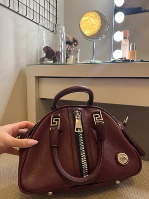 Vinröd handväska  - Snygg vinröd handväska i läder med silverfärgade metalldetaljer och dekorativa mönster på handtagen. Väskan har två handtag, dragkedja framtill och rundad form. Perfekt för dig som vill ha en unik accessoar.