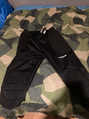 Svarta målvaktsbyxor från Uhlsport - Svarta målvaktsbyxor från Uhlsport med vadderade skydd på knän och höfter. Byxorna har resår i midjan med snörning och är tillverkade i ett slitstarkt syntetmaterial. Perfekta för fotbollsmålvakter som vill ha extra skydd under match och träning. Kan gå lägre.