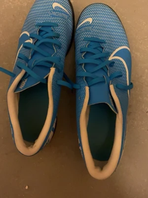 Blå Nike fotbollsskor med vit detalj - Snygga blå fotbollsskor från Nike med vita detaljer och svart sula. Skorna har snörning och texten 'JUST DO IT' på sidan. Ovandelen är i syntetmaterial med mönstrad yta för bättre bollkontroll. Perfekta för inomhusspel.