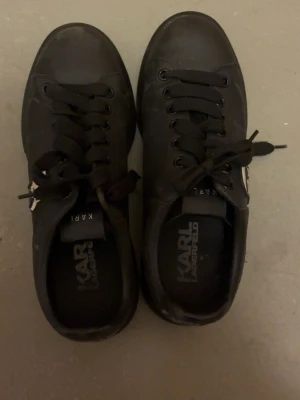 Svarta sneakers Karl Lagerfeld - Snygga helsvarta sneakers från Karl Lagerfeld med svart snörning och en cool logga i vitt och svart på sidan. Skorna är låga och har en stilren design i slätt lädermaterial. Perfekta för dig som gillar minimalistisk och trendig stil.