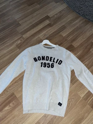 Ljusgrå tröja från Bondelid - Ljusgrå långärmad tröja från Bondelid med svart broderad text 'BONDELID 1956' på bröstet. Tröjan har rund halsringning och ribbade muddar vid ärmslut och nederkant. Perfekt för en avslappnad och stilren look.