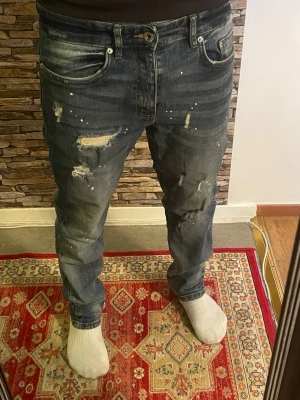 Mörkblå slitna jeans med detaljer - Säljer ett par mörkblå jeans med distressed detaljer och färgstänk. Jeansen har en relaxed fit och klassisk femficksmodell. Materialet är denim i bomull och de har slitningar och hål för en trendig, avslappnad look.