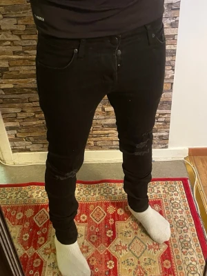 Svarta skinny jeans med slitningar - Säljer ett par svarta skinny jeans med slitningar vid knäna. Jeansen har klassisk femficksdesign och är i stretchigt material som sitter tight längs benen. Perfekta för en trendig och avslappnad stil.