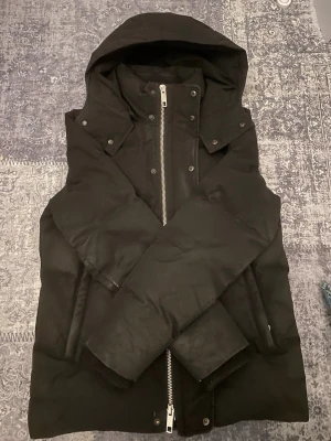 Svart pufferjacka med huva - Snygg svart pufferjacka med stor huva och kraftig dragkedja framtill. Jackan har en stilren design med tryckknappar vid kragen och breda, vadderade ärmar. Perfekt för kalla dagar när du vill hålla värmen och ändå se cool ut.