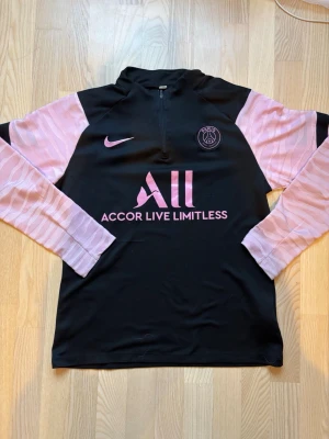 PSG träningströja svart/rosa Nike - Snygg långärmad träningströja från Paris Saint-Germain i svart med rosa detaljer och logga. Tröjan har halv dragkedja, rosa mönstrade ärmar och tryckt sponsorlogga på bröstet. Perfekt för fotbollsträning eller sportiga outfits. Har lite skador