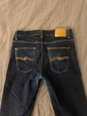 Mörkblå jeans från Nudie Jeans - Snygga mörkblå jeans med fet wash från Nudie Jeans med klassisk femficksmodell och kontrastsömmar. Bakfickorna har Nudies ikoniska broderi i orange. Jeansen har normal midja och raka ben. Perfekta för en stilren och avslappnad look. Seriösa bud kan godkännas.