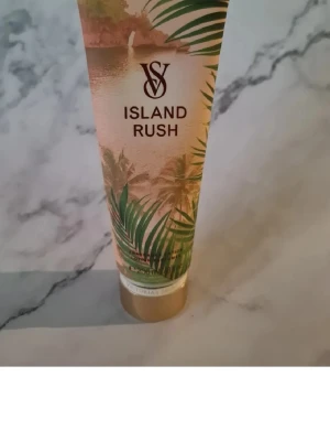 Victoria's Secret Island Rush lotion - Island Rush lotion från Victoria's Secret med tropisk doft av kokos, tahitisk gardenia och tränoter. Tuben har ett snyggt designat motiv med palmer och guldiga detaljer. Perfekt för dig som vill ha en exotisk och fräsch känsla på huden. Helt ny och oöppnad