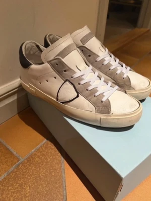 Vit-gråa sneakers från Philippe Model - Snygga sneakers från Philippe Model i vitt läder med grå mockadetaljer och svart hälparti. Klassisk låg modell med vita skosnören och den ikoniska skölden på sidan. Perfekta för en stilren och trendig look.