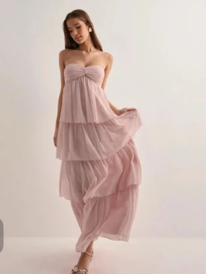 Ljusrosa  med volanger - Säljer en romantisk ljusrosa långklänning med flera lager volanger och omlott över bysten. Klänningen är axelbandslös och har en luftig, flowy design som passar perfekt för sommarens alla tillfällen. Använd en gång så i väldigt fint skick. 