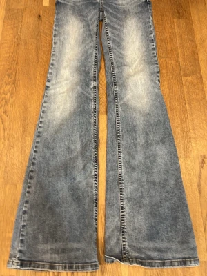 Blå utsvängda jeans med fickdetalj - Blå jeans med utsvängda ben och snygg slitning. Jeansen har en bakficka med  knapp och dekorativ vit söm. Dom är från Nelly, storlek 34. Dom har ett väldigt litet hål lägnst ner men de e inte synligt. 