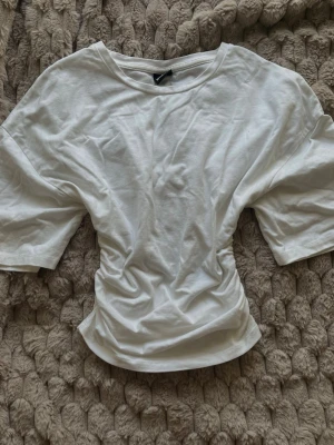 Vit t-shirt med scrunch på sidorna - Gina Tricot - Jättefin vit t-shirt från Gina Tricot i storlek XS. T-shirten har rund halsringning och korta ärmar. Även scrunch på sidorna som ger en timglas effekt. Tillverkad i mjuk bomull, perfekt för en enkel och clean look. Funkar i alla väder. Oanvänd.