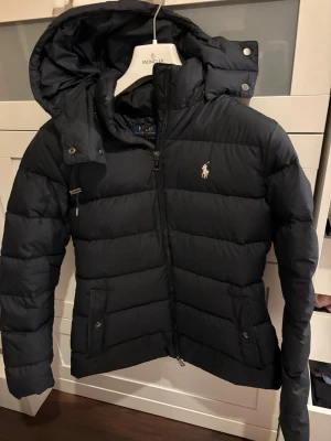Svart dunjacka från Polo Ralph Lauren - Snygg svart dunjacka från Polo Ralph Lauren med avtagbar huva, dragkedja framtill och två fickor med knappstängning. Jackan har det klassiska Polo-logotypen broderad på bröstet och är perfekt för kalla vinterdagar. Fin feminin passform, storlek M men passar även S bra. Sparsamt använd och i gott skick!