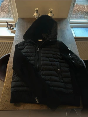 Svart Moncler Cardigan  - Snygg svart pufferjacka från Moncler med stickade ärmar och huva. Jackan har dragkedja framtill, fickor med dragkedja och en extra ficka på ärmen. Perfekt för kyliga dagar. Den är i väldigt fint skick efter som jag har tagit väll hand om den. Jag köpte den på NK i Gbg för nått år sen men har tyvärr slarvat bort kvittot. Den är i storlek L men passar även den som bär vanliga viss storlek M. 