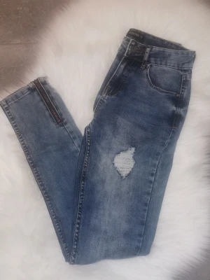 Blå slitna jeans med dragkedjor, storlek 34 - Säljer ett par blå jeans från Foster med slitningar på låren och snygga dragkedjor vid bensluten. Jeansen har klassisk femficksmodell och är tillverkade i denim med en tvättad look. Perfekta för en avslappnad och trendig stil. Köpta utomlands. Storlek 34. Är knappt använd, jättefin skick. 