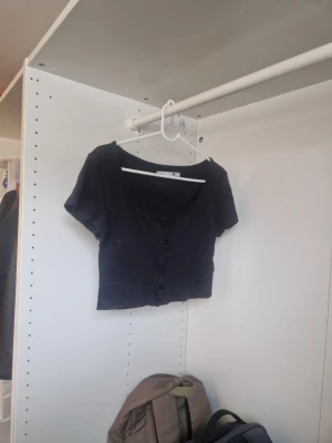 Svart croppad topp med knappar - En svart croppad topp med korta ärmar och knappar framtill. Toppen har en djup rundringning och är gjord i ett ribbat material som ger en snygg och bekväm passform. Perfekt att styla med höga jeans eller kjol.