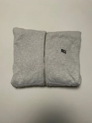 Grå zip hoodie från lexington  - Snygg grå hoodie med dragkedja från lexington. Klassisk design med liten logga på bröstet, huva och två fickor framtill. Tillverkad i mjukt bomullsmaterial som känns skönt mot huden. Perfekt för en avslappnad och stilren look.