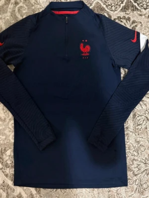 Frankrike tröja - Mörkblå långärmad träningströja från Nike med Frankrikes landslagsmärke och röd Nike-logga på ärmarna. Tröjan har halv dragkedja framtill och är tillverkad i ett lätt, stretchigt material som passar perfekt till fotbollsträning. Storlek S.