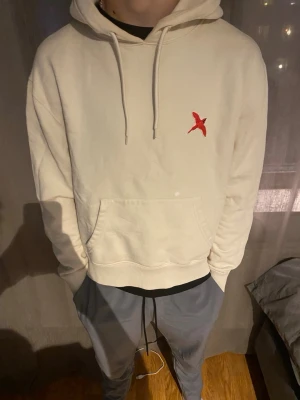 Vit hoodie med röd fågel - Snygg vit hoodie med huva och känguruficka framtill. På bröstet finns en broderad röd fågel som detalj. Hoodien har dragsko i huvan och ribbade muddar vid ärmslut och nederkant. Perfekt för en chill och avslappnad stil.
