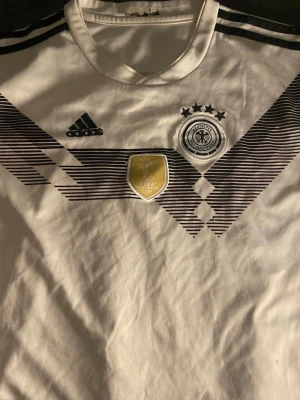 Tysklands fotbollströja Adidas - Vit Tysklands landslagströja från Adidas med svarta detaljer och mönster på bröstet. Broderad landslagsemblem, tre stjärnor och FIFA-märke framtill. Namnet 'Neuer' tryckt på ryggen. Perfekt för fotbollsfans!