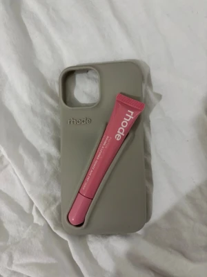 Rhode mobil skal med läppglans  - Snyggt grått/beiget mobilskal från Rhode med fack för läppglans till iPhone 15. Skalet är gjort i mjukt silikonmaterial och har en stilren design med Rhode-loggan på baksidan. Perfekt för dig som vill ha både skydd och funktion i ett. Läppglanset har tappat lite av färgen på texten, utöver det är skalet helt oanvänt.❤️🌸