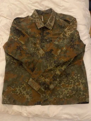 Camouflage overshirt i militärstil - Säljer en overshirt i klassiskt militärinspirerat camouflagemönster med gröna, bruna och svarta toner. Skjortjackan har krage, knappstängning framtill och en bröstficka. Perfekt för dig som gillar streetwear och utility-vibe.