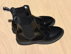 Svarta Chelsea boots från Dr. Martens - Snygga svarta Chelsea boots från Dr. Martens i blankt läder med klassiska elastiska paneler på sidorna och dragflik baktill. Skorna har en rundad tå och robust sula, perfekt för en cool och stilren look.