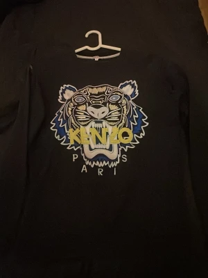 Svart Kenzo hoodie med tigertryck - Svart hoodie från Kenzo med ikoniskt tigertryck i blått, vitt och gult på bröstet samt texten 'KENZO PARIS'. Hoodien har långa ärmar och en klassisk passform. Perfekt för dig som vill sticka ut med en streetstyle-look.