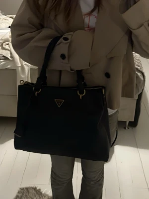 Svart handväska från Guess - Säljer en snygg svart handväska från Guess med klassisk trekantslogga i metall på framsidan. Väskan har guldfärgade detaljer, två handtag och en justerbar axelrem. Materialet är slätt och lyxigt läder, perfekt för dig som gillar stilrena accessoarer. Har två stora fack, med andra små fack i. 