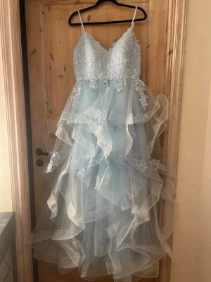 Ljusblå balklännning - Ljusblå långklänning med v-ringad spetsöverdel och tunna axelband. Kjolen har flera lager av volangigt tyll och spetsdetaljer som ger ett drömmigt och elegant intryck. Perfekt för student eller bal.