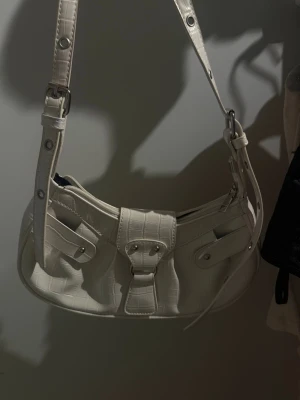 Vit axelremsväska i croco-mönster - Snygg vit handväska med croco-mönstrad yta och justerbar axelrem. Väskan har två fickor framtill med lock och knappar samt en dragkedja upptill. 