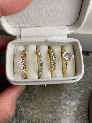 Fyra guld pläterade ringar i guld 18k med stenar - Säljer ett set med fyra eleganta guldringar, alla prydda med gnistrande stenar i olika former. En ring har en stor rund klar sten, medan de andra har mindre stenar i rad, både runda och avlånga. Perfekta för att mixa och matcha för en trendig look.